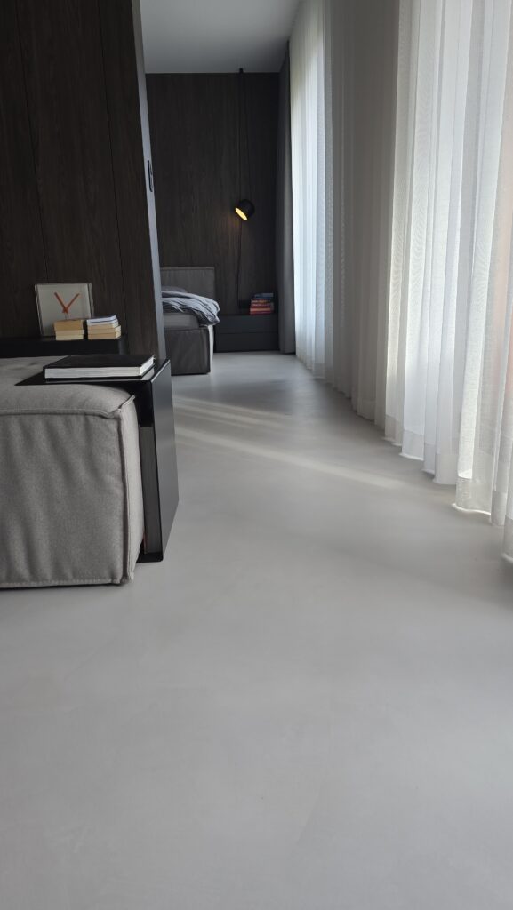 Podłoga z szarego mikrocementu Festfloor w nowoczesnej sypialni z ciemnymi ścianami i jasnymi zasłonami